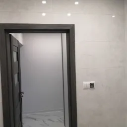 Wykończona łazienka z jasnymi płytkami imitującymi kamień i otwartymi, ciemnoszarymi drzwiami z przeszkleniami, widoczne gniazdko elektryczne i włącznik światła na ścianie.