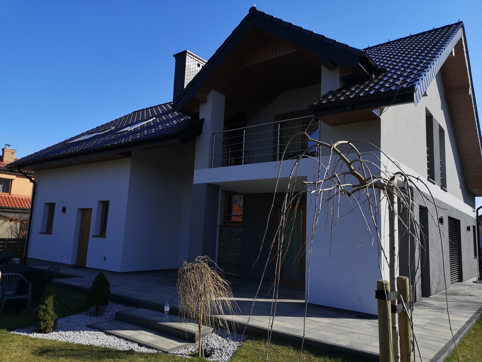 Nowoczesny dom jednorodzinny z balkonem i ciemną dachówką, jasna elewacja, szare wykończenia, zadbany ogród z kamieniami i niewielkimi drzewkami.