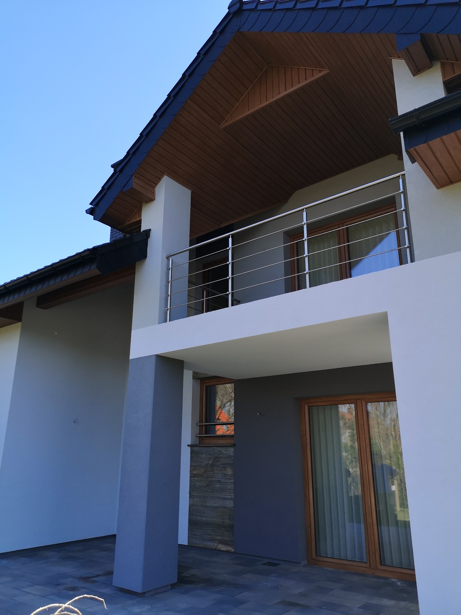 Nowoczesny dom z jasną elewacją, balkonem ze stalową balustradą i drewnianą podbitką dachu, widok z dołu na błękitne niebo.