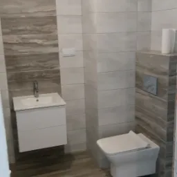 Wykończona łazienka z płytkami imitującymi drewno na podłodze i ścianie, wisząca toaleta z przyciskiem spłukującym oraz biała szafka z umywalką i baterią.