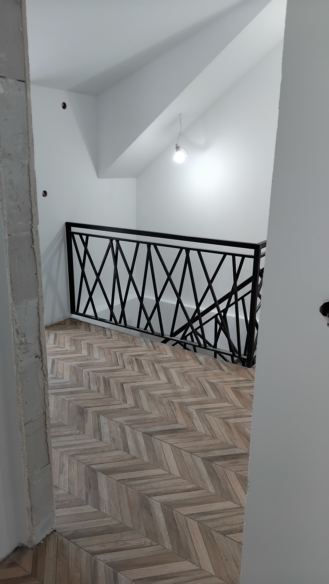 Ukończone schody z drewnopodobną podłogą ułożoną w jodełkę i czarną, metalową balustradą o geometrycznym wzorze, widoczne z góry, w jasnym wnętrzu.