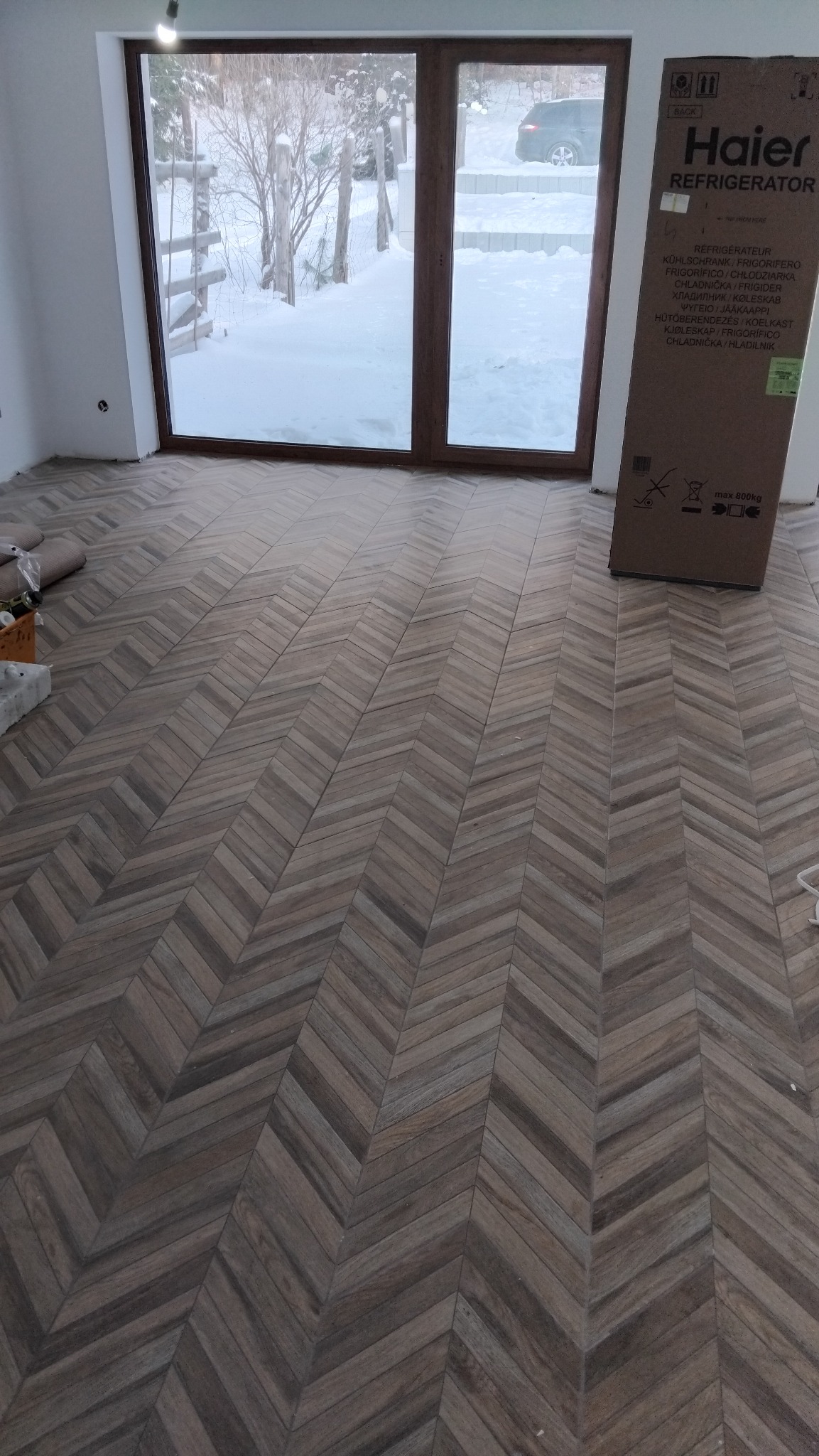 Ukończona podłoga z płytek ułożonych w jodełkę, widok na okno z zimowym krajobrazem i karton po lodówce. Jasne wnętrze, naturalne światło.