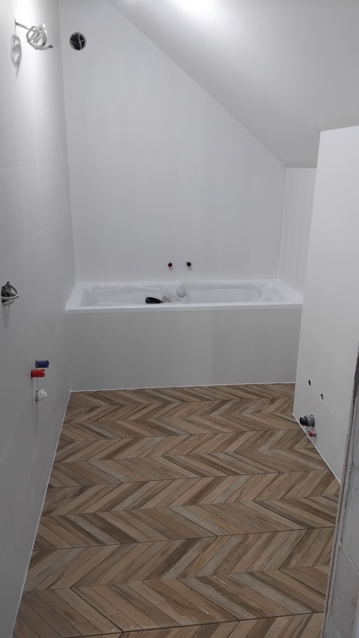 Biała łazienka na poddaszu z drewnopodobną podłogą ułożoną w jodełkę. Wanna obudowana płytkami, widoczne przyłącza wodne. Świeżo położone płytki na ścianach.