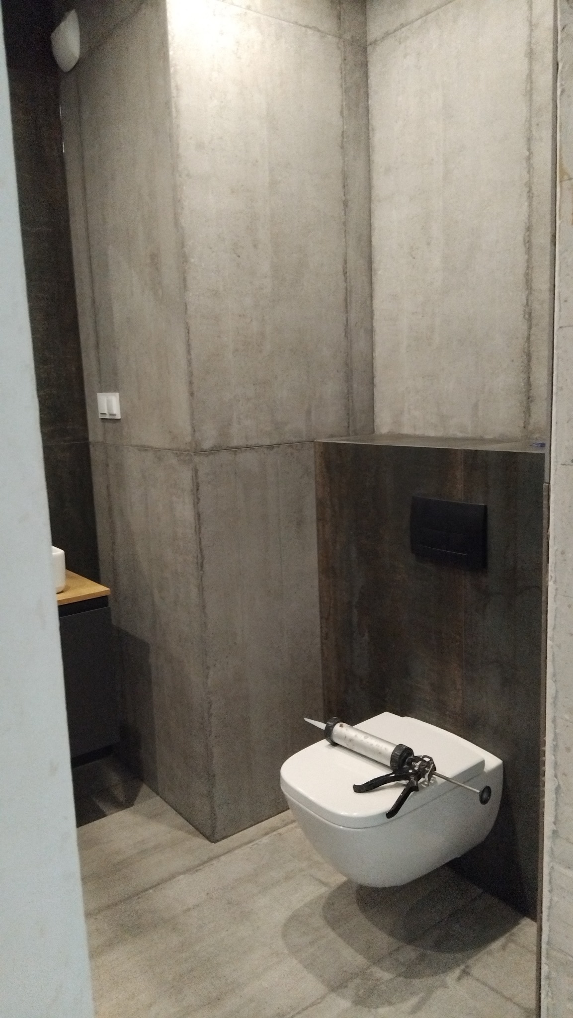 Nowoczesna łazienka z betonowymi ścianami i podwieszaną toaletą. Na sedesie leży pistolet do silikonu, sugerujący prace wykończeniowe.