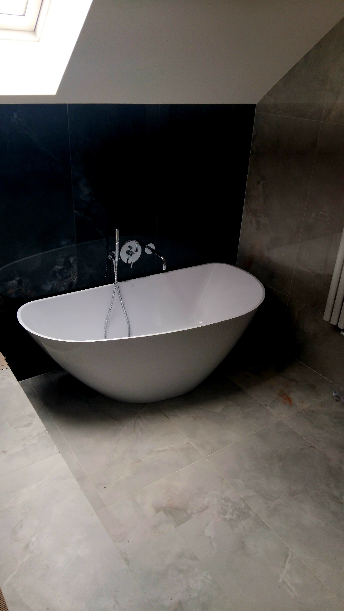 Nowoczesna, wolnostojąca wanna w łazience z czarną ścianą i marmurową podłogą. Widoczne okno dachowe i chromowana armatura.