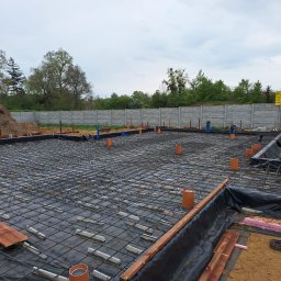 MAX-BUD - Przygotowanie fundamentów pod dom murowany: zbrojenie, izolacja folią i rury instalacyjne na czarnej membranie, w tle betonowy płot i kopiec ziemi.