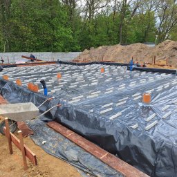 MAX-BUD - Przygotowanie fundamentów pod dom: folia izolacyjna, stalowe zbrojenie i betonowe podkładki dystansowe na placu budowy, w tle pracownicy i hałda ziemi.