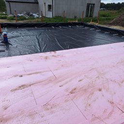 MAX-BUD - Przygotowanie podłoża pod płytę fundamentową: różowy styropian ułożony na folii hydroizolacyjnej w wykopie, widoczne drewniane szalunki i fragment budynku w tle.