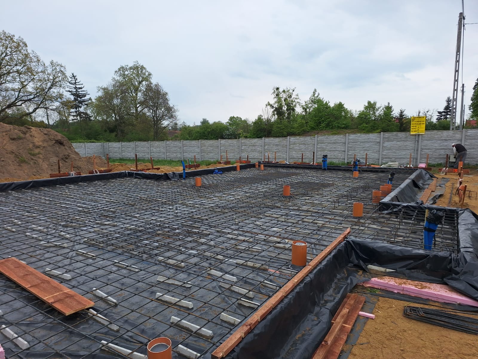 Przygotowanie fundamentów pod dom murowany: zbrojenie, izolacja folią i rury instalacyjne na czarnej membranie, w tle betonowy płot i kopiec ziemi.