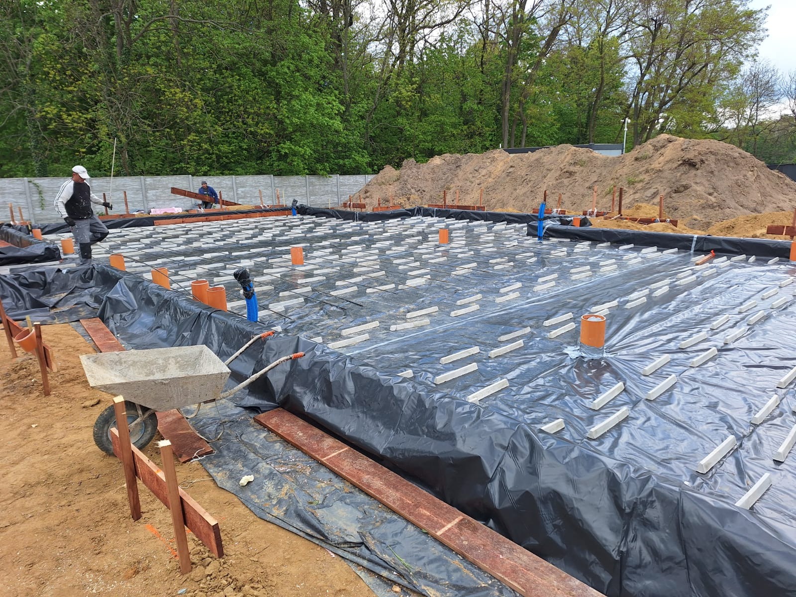 Przygotowanie fundamentów pod dom: folia izolacyjna, stalowe zbrojenie i betonowe podkładki dystansowe na placu budowy, w tle pracownicy i hałda ziemi.