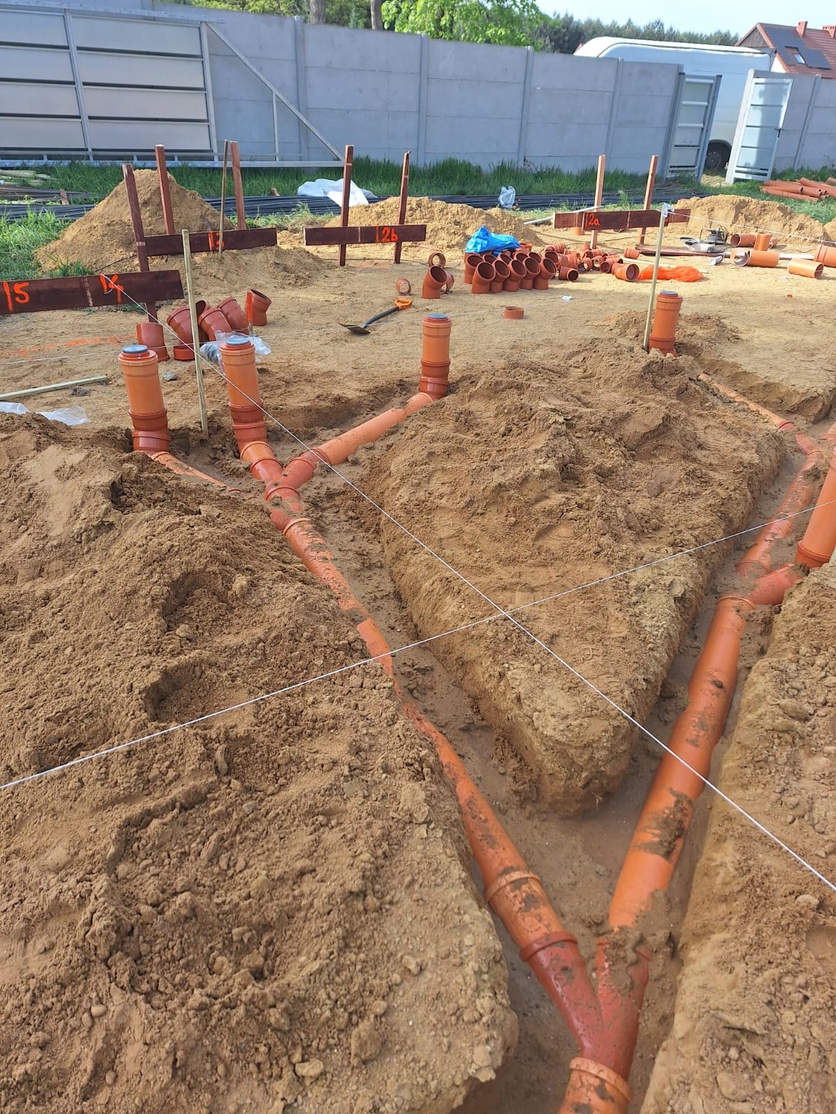 Instalacja pomarańczowych rur kanalizacyjnych w wykopie w gruncie z widocznymi deskami pomiarowymi i zapasem rur na powierzchni.
