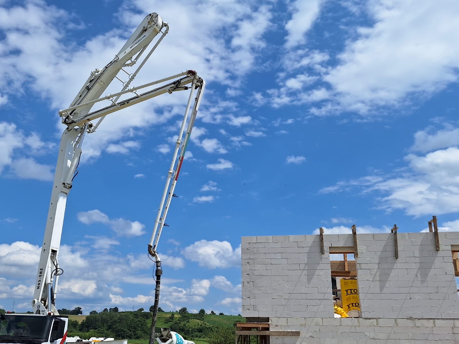 Betonomieszarka z wysięgnikiem pompuje beton do wznoszonej ściany budynku z białych bloczków na tle błękitnego nieba z obłokami.