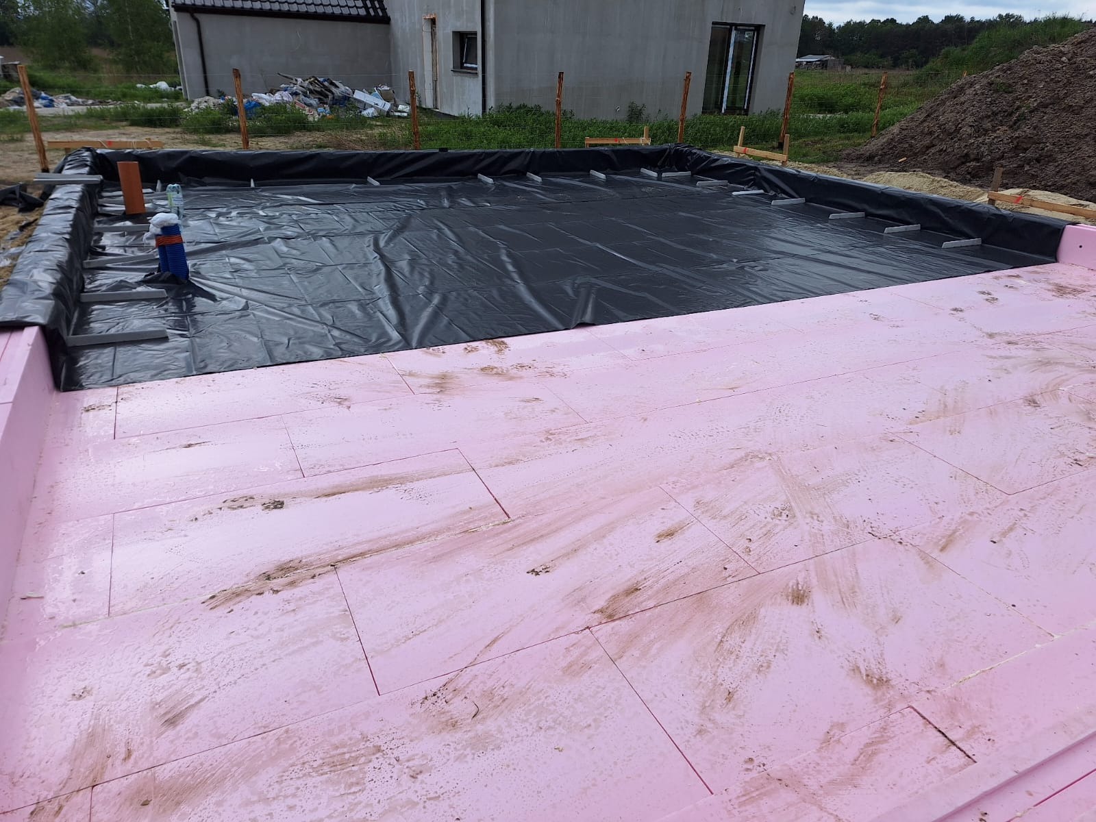 Przygotowanie podłoża pod płytę fundamentową: różowy styropian ułożony na folii hydroizolacyjnej w wykopie, widoczne drewniane szalunki i fragment budynku w tle.