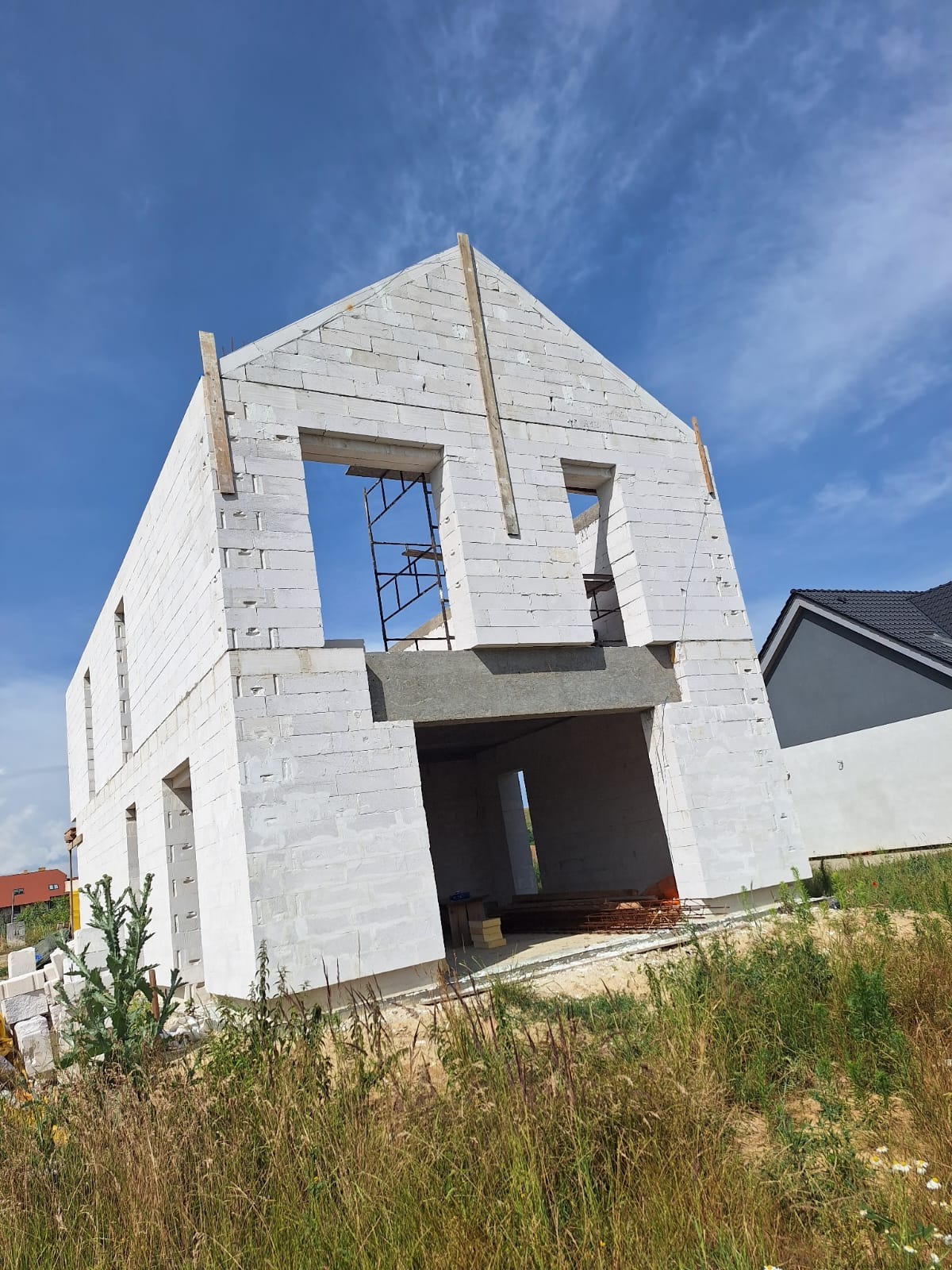 Niedokończony dwukondygnacyjny dom z białego bloczka, widoczne rusztowanie w otworze okiennym, drewniane stemple podtrzymujące szczyt, w tle fragment dachu drugiego budynku.