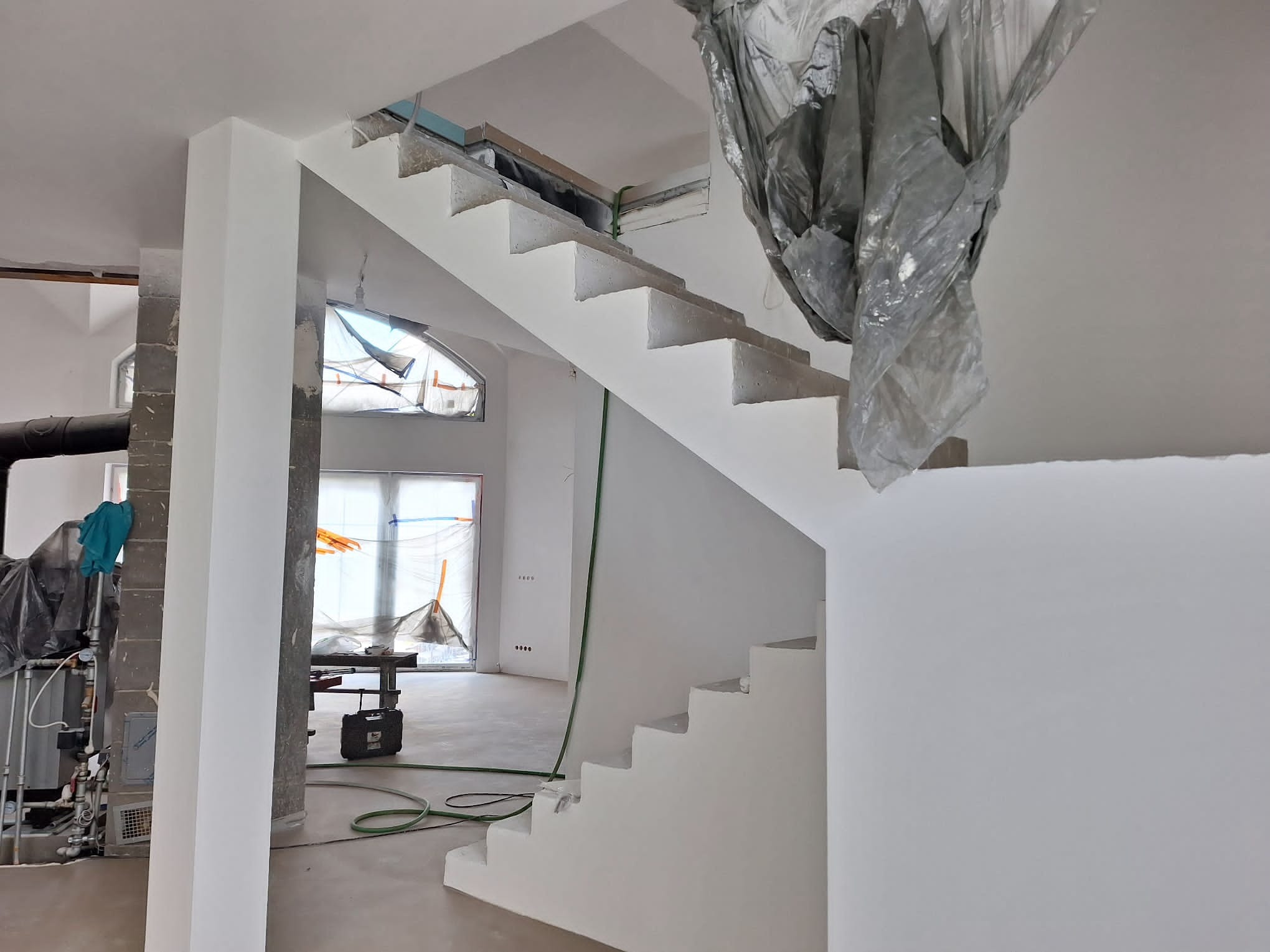 Wnętrze domu w trakcie prac wykończeniowych: białe, surowe schody bez balustrady, zabezpieczone folią, widoczne narzędzia i sprzęt budowlany w tle, okno z folią ochronną.