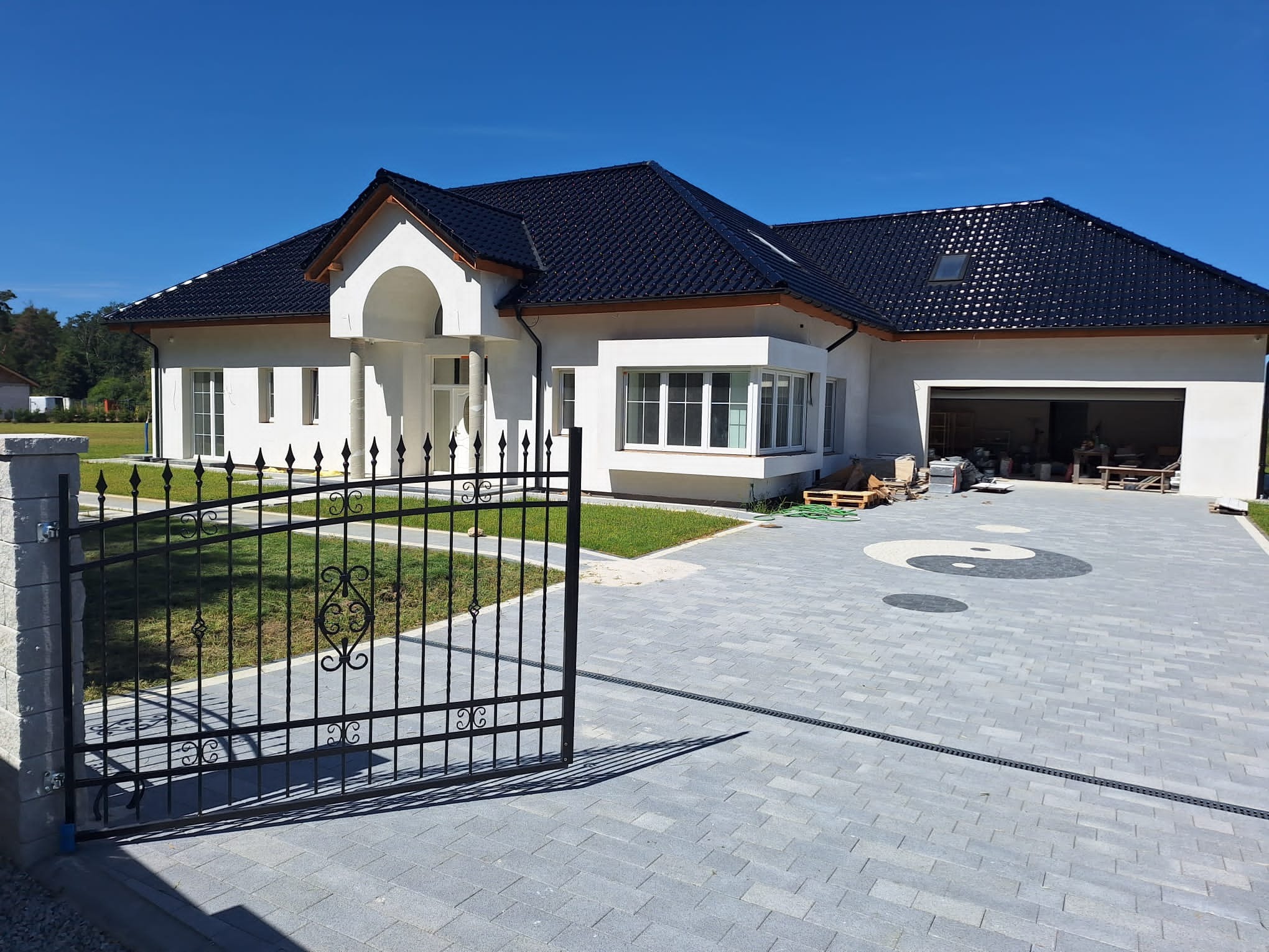 Nowoczesny dom jednorodzinny z czarnym dachem i białymi ścianami, otoczony trawnikiem i wybrukowanym podjazdem z motywem Yin Yang, widok zza otwartej, metalowej bramy.