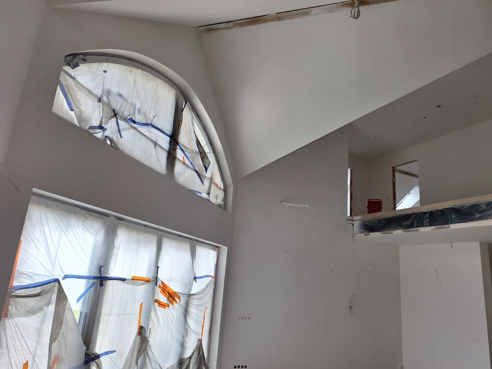 Wnętrze w trakcie prac wykończeniowych: zabezpieczone folią okna o nietypowym kształcie, świeżo pomalowane ściany na wysoki połysk, widoczne niedokończone instalacje elektryczne.