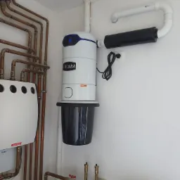 Biały system centralnego odkurzania BEAM zamontowany na ścianie w pomieszczeniu technicznym, widoczne rury instalacji grzewczej i inne urządzenia.