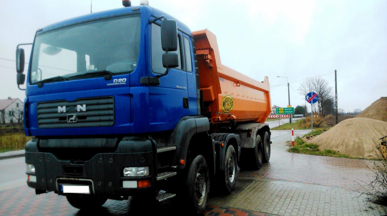 Niebiesko-pomarańczowa wywrotka MAN TGA 41.430 8x6 na mokrej kostce brukowej, w tle sterta piasku i znak drogowy.