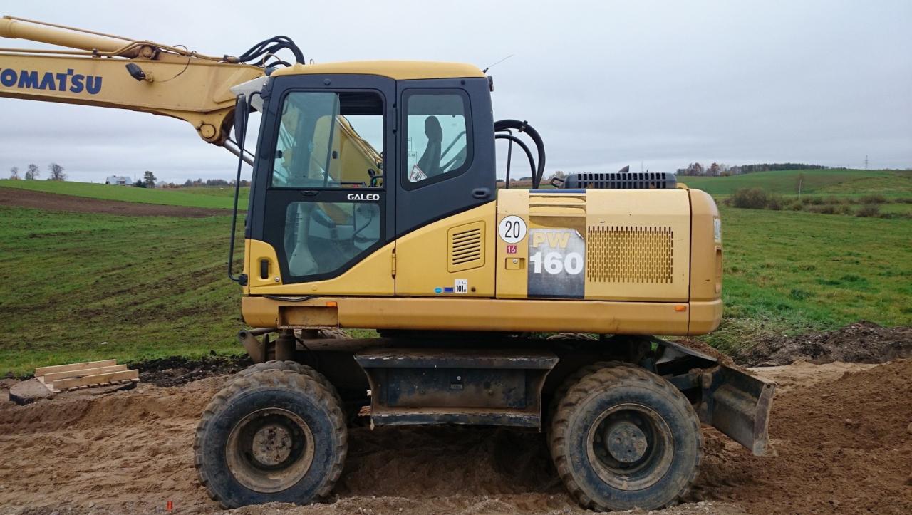Żółta koparka kołowa Komatsu PW160ES-7K stoi na ziemi z widocznym ramieniem i kabiną operatora, w tle zielone pole i pochmurne niebo.
