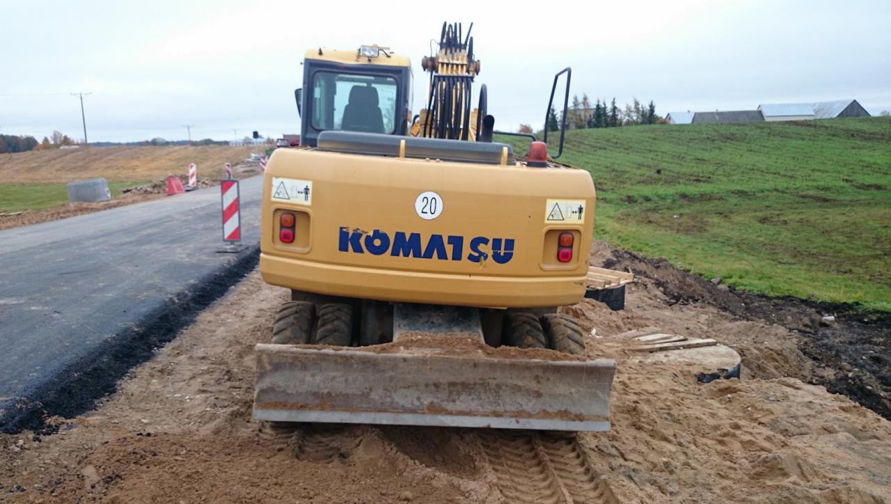 Żółta maszyna Komatsu PW160ES-7K, widok z tyłu, stoi na nierównym, piaszczystym terenie obok nowo budowanej drogi z widocznymi znakami ostrzegawczymi i zielonym wzgórzem w tle.