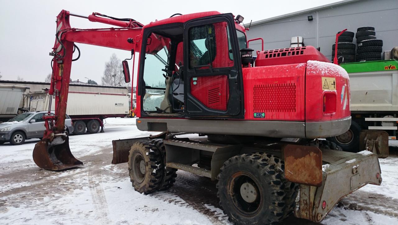 Czerwona koparka kołowa Komatsu PW140-7 na ośnieżonym placu, widoczna z boku, z otwartą kabiną i łyżką opuszczoną do ziemi, w tle zaparkowane samochody i naczepy.