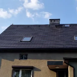 Dom z dachem pokrytym ciemnobrązową dachówką ceramiczną, z trzema oknami dachowymi i kominem z cegły, sąsiadujący z budynkiem z czerwonym dachem z blachy falistej na tle błękitnego nieba z chmurami.