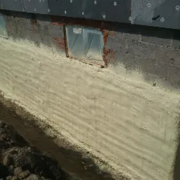 Ocieplona pianą PUR ściana fundamentowa budynku z widocznymi otworami okiennymi zabezpieczonymi folią. Widoczny fragment ocieplenia ściany powyżej.