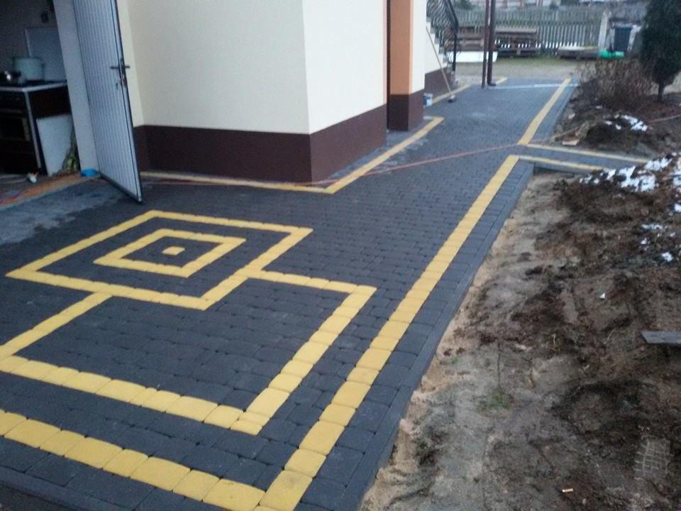 Ułożona, ciemnoszara kostka brukowa z żółtymi akcentami tworzącymi geometryczny wzór przed budynkiem. Częściowo widoczna ziemia z resztkami śniegu po prawej stronie.