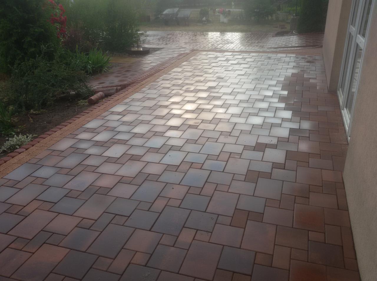 Ułożona kostka brukowa w odcieniach brązu i szarości, tworząca wzór na podwórku przy budynku z oknem, z obrzeżem z drobnej kostki i widoczną roślinnością w tle.