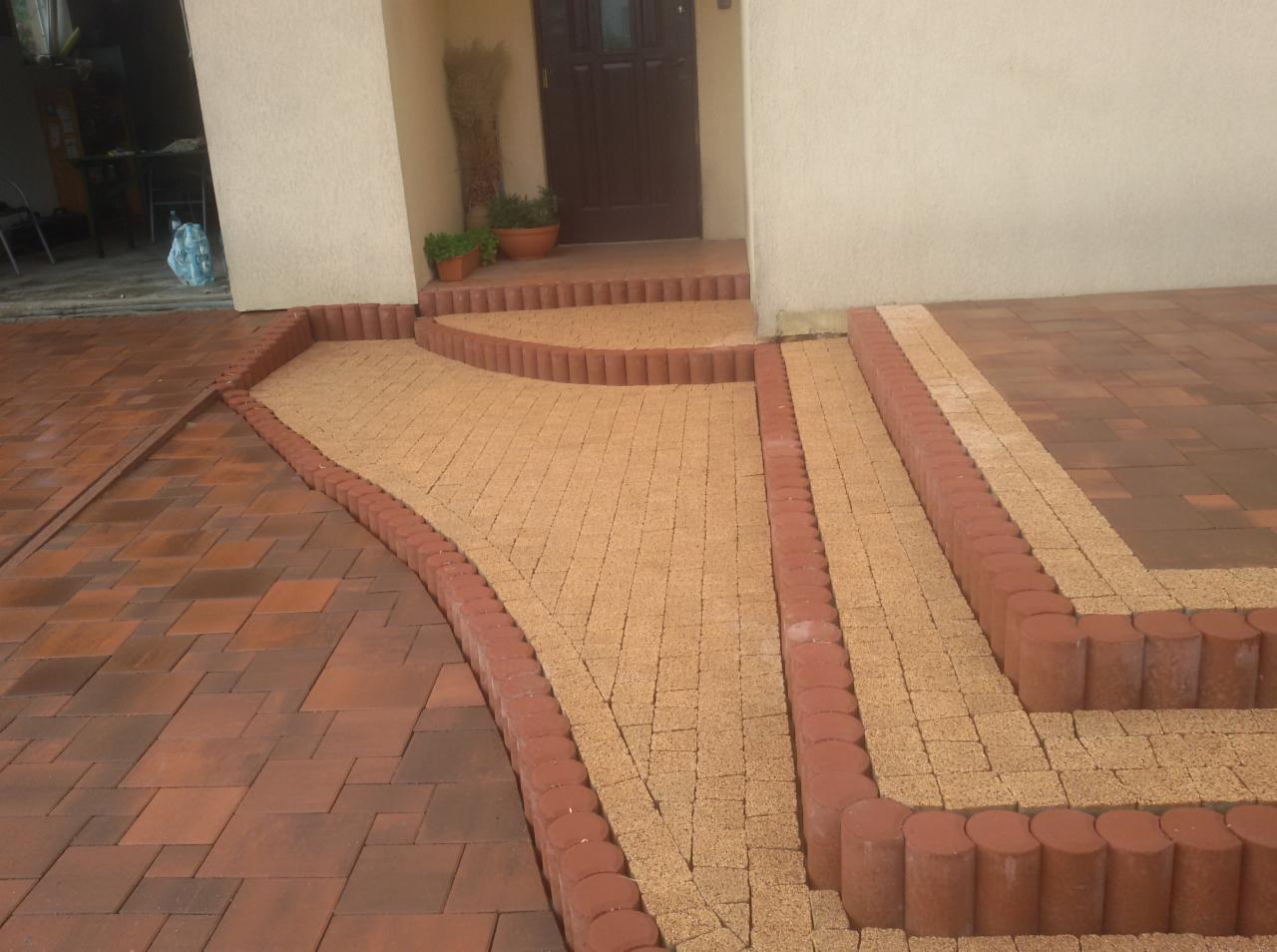 Ułożona nawierzchnia z kostki brukowej w dwóch kolorach: beżowej i ceglanej, obramowana okrągłymi obrzeżami, prowadząca do wejścia domu z roślinami w donicach.