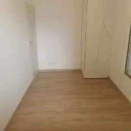 Pokój połóżony panele podłogowe w odcieniach jasnego drewna, widoczne białe ściany, białe drzwi oraz biała szafa z wystającym kablem, gniazdka elektryczne na ścianie.