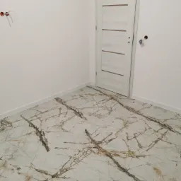 Wykończona podłoga z płytek imitujących marmur w jasnych odcieniach, widoczna biała listwa przypodłogowa, białe ściany i białe drzwi z poziomymi wstawkami w nowym mieszkaniu, puszki elektryczne bez...