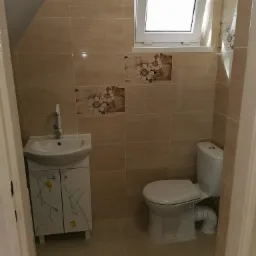 Ukończona łazienka na poddaszu z beżowymi płytkami imitującymi marmur, białą umywalką z szafką, toaletą i oknem dachowym.