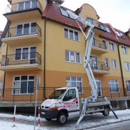 Technikal Ernest Kalus - Podnośnik koszowy z wysięgnikiem opartym o dach budynku. Biały pojazd na tle żółtej elewacji, zima, śnieg na chodniku. Widoczny klimatyzator na dachu.