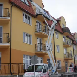 Podnośnik koszowy CTE Z 17L na tle budynku z balkonami. Żółto-pomarańczowa elewacja, czerwony dach. Zima, śnieg na chodniku. Auto z napisem 'Podnośnik'.
