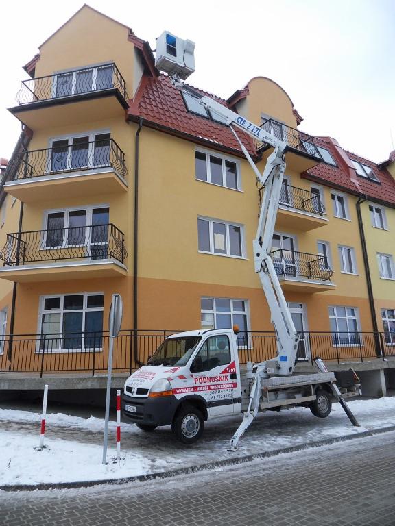 Podnośnik koszowy z wysięgnikiem opartym o dach budynku. Biały pojazd na tle żółtej elewacji, zima, śnieg na chodniku. Widoczny klimatyzator na dachu.