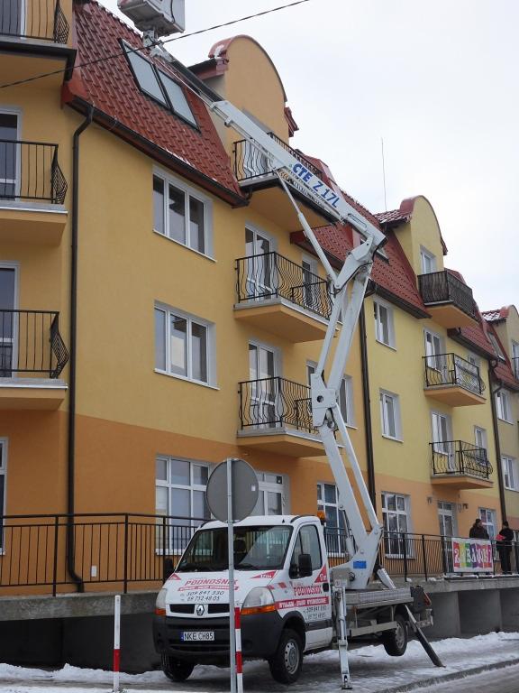 Podnośnik koszowy CTE Z 17L na tle budynku z balkonami. Żółto-pomarańczowa elewacja, czerwony dach. Zima, śnieg na chodniku. Auto z napisem 'Podnośnik'.