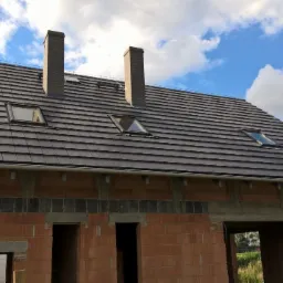 Dom w budowie z ciemną dachówką, widoczne kominy, okna dachowe i rynny. Ściany z czerwonej cegły w trakcie wznoszenia, błękitne niebo w tle.