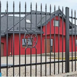 Czarne, metalowe ogrodzenie panelowe z dekoracyjnymi elementami na tle czerwonego domu z szarym dachem. Widoczne pionowe pręty i ozdobne motywy w kształcie elips.
