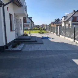 Ułożona szara kostka brukowa przed nowoczesnym domem z elementami z cegły, widoczne schody wejściowe i zadbany trawnik z ozdobnymi krzewami w tle.