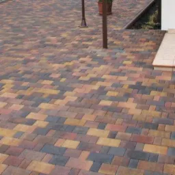 Kolorowa kostka brukowa ułożona we wzór jodełki, prowadząca do schodów z donicą, oświetlona naturalnym światłem.