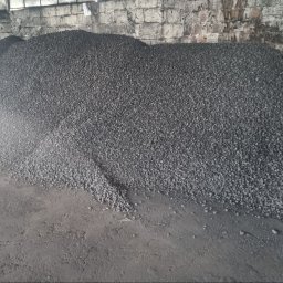 El-Agro Elżbieta Faber - Duży pryzmat czarnego ekogroszku na betonowej posadzce wewnątrz magazynu, z widocznym workiem w tle i fragmentem murowanej ściany.