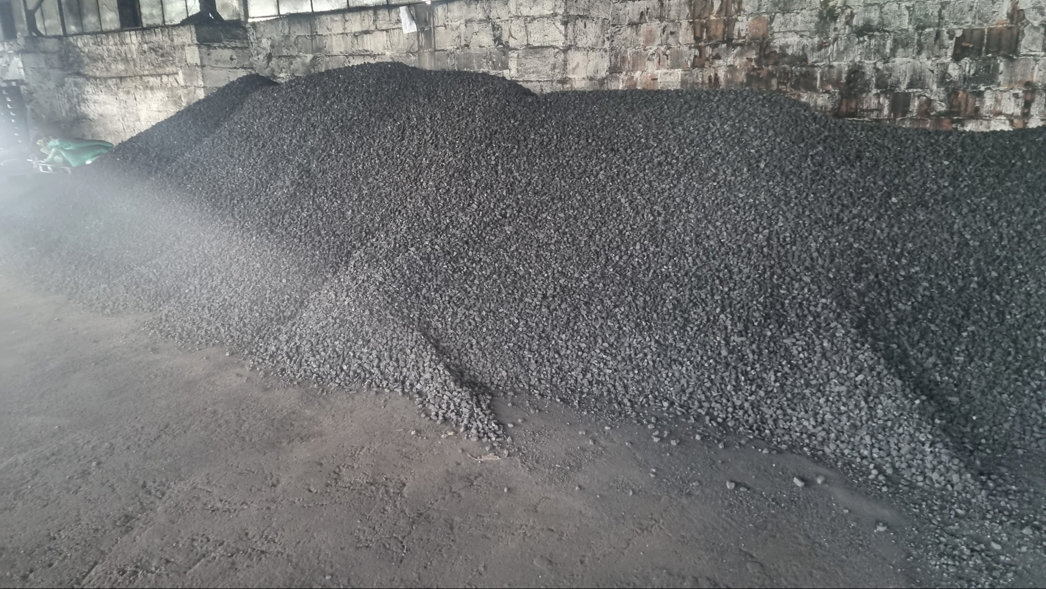 Duży pryzmat czarnego ekogroszku na betonowej posadzce wewnątrz magazynu, z widocznym workiem w tle i fragmentem murowanej ściany.