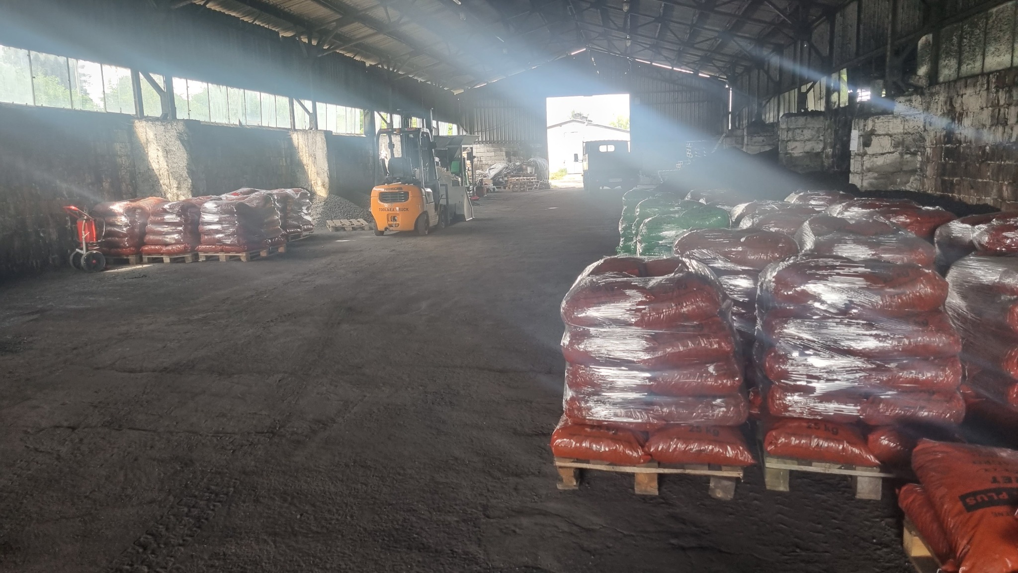 Magazyn z workami opału na paletach, w tle wózek widłowy i wpadające światło słoneczne przez okna dachu.