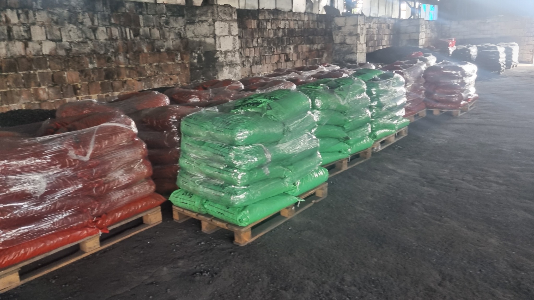 Magazyn z rzędami palet z workami węgla, część worków czerwona, część zielona, zapakowane w folię, tło stanowi ceglana ściana.