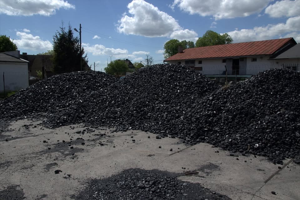 Góra ekogroszku na placu składowym w słoneczny dzień. Widoczny budynek w tle i fragment nieba z chmurami. Materiał opałowy przygotowany do sprzedaży.