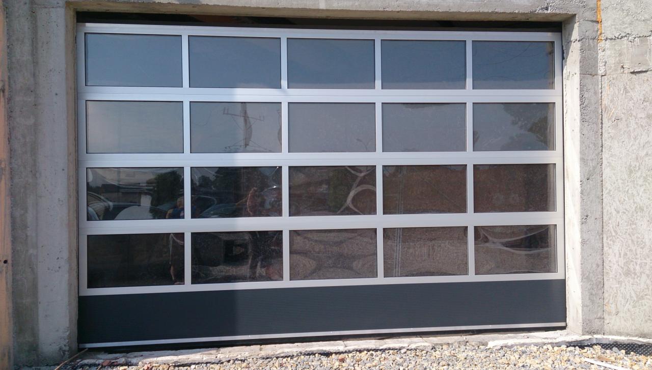 Zamknięta, segmentowa roleta garażowa z aluminiową ramą i szybami, widoczna od zewnątrz, z szarym panelem dolnym i betonową wnęką.