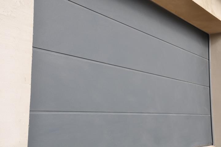 Szara, segmentowa brama garażowa z poziomymi przetłoczeniami, zamontowana w jasnym murze. Widok z bliska, ukazujący detale konstrukcji i fakturę materiału.