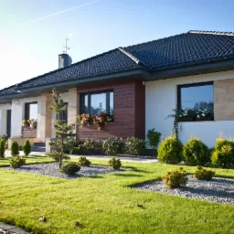 Nowoczesny dom jednorodzinny z zadbanym ogrodem: trawnik, rabaty z krzewami i drzewkami, okna z drewnianymi obudowami i kwiatami, jasna elewacja z elementami dekoracyjnymi.